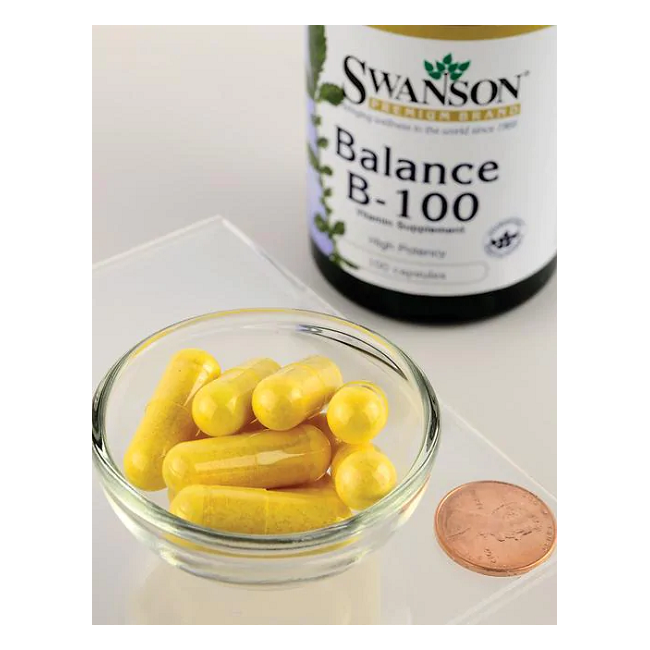 Next to a penny and a bottle labeled "Swanson Balance Vitamin B-100 Complex," a glass bowl holds yellow capsules rich in B vitamins.