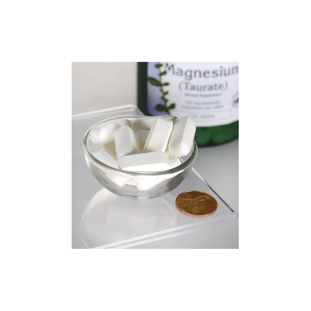 Un frasco de Swanson Taurato de Magnesio 100 mg 120 tab sentado junto a un cuenco de pastillas blancas.