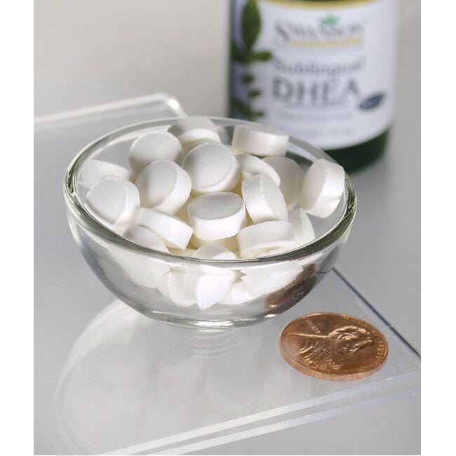 Un bol de pastillas blancas junto a un frasco de Swanson DHEA - 25 mg 60 pastillas Sabor Cereza.