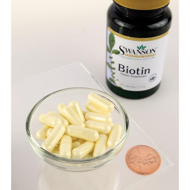 Un frasco de suplemento dietético de Swanson Biotin - 5 mg 100 cápsulas junto a un penique sobre una mesa.